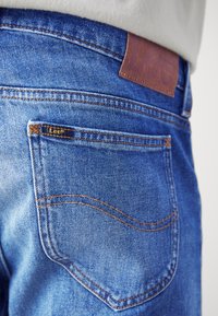 Blue denim jeans featuring a brown leather patch at the waist and a small brand tag on the back pocket with orange stitching details.