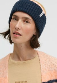 Knitted beanie in navy with pink and tan stripes, featuring a ribbed texture. The individual wears a peach cardigan and a light tan shirt beneath.
