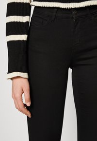 Czarne skinny jeans o gładkiej fakturze, wysoki stan i smukły krój. Ręka opiera się na biodrze, podkreślając detale szwów.