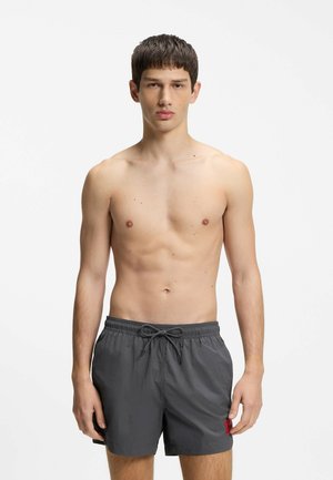 Jeune homme debout, face à l'avant, torse nu, portant un short de bain gris foncé à cordon contre un fond clair uni.