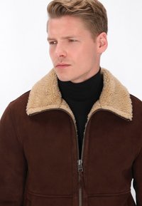 Jachetă din suede maro, cu guler din blană bej, închidere cu fermoar și detaliu cu buton. Prezintă o textură netedă și un design ajustat.