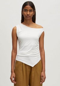 Asymmetrische witte gebreide top met een enkele schouderband, voorzien van geplooide textuur en een puntige zoom, gecombineerd met lichtbruine broeken.