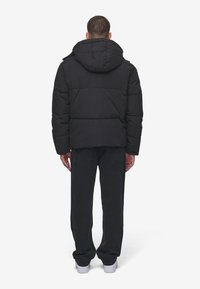 Svart pufferjakke med hette, med et quiltet design og droppede skuldre. Båret over mørke jeans og hvite joggesko.