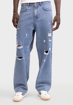 Mann kledd i løstsittende blå revne jeans og hvite joggesko med svarte striper, står mot en ensfarget lys bakgrunn.