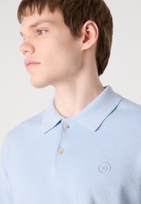 Gaiši zila polo krekls, teksturēta auduma, īsām piedurknēm, ar diviem pogām, izšūta logo kreisajā krūtī, klasiskā apkakles dizains.