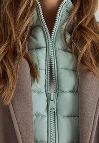 Hellblau gestepptes Weste, die unter einem braunen Mantel getragen wird. Sie verfügt über einen durchgehenden Reißverschluss mit passendem Zipper und strukturierten Mustern auf der Weste.