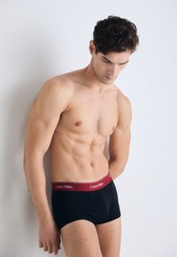 Zwarte herenslips met een brede bordeauxrode tailleband waarop het logo "Calvin Klein" staat. Gladde stof met een aansluitend ontwerp.