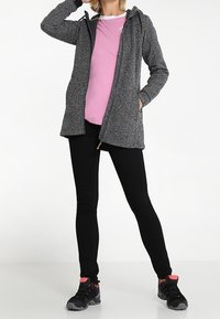 Kvinders grå, struktureret lang jakke med hætte, kombineret med en lyserød skjorte, sorte leggings og sorte sneakers med lyserøde accenter.