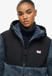 Giacca nera e blu con una texture morbida e pelosa sulle maniche e nella parte inferiore, dotata di zip frontale e patch con logo sul petto.