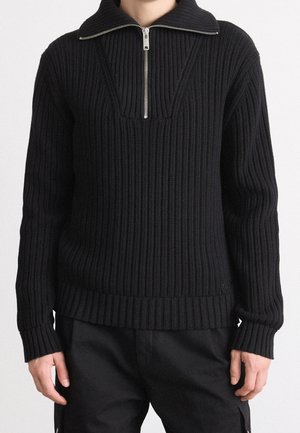 Person trägt schwarzen gerippten Strickpullover mit halbem Reißverschluss am Kragen und langen Ärmeln, kombiniert mit schwarzen Hosen.
