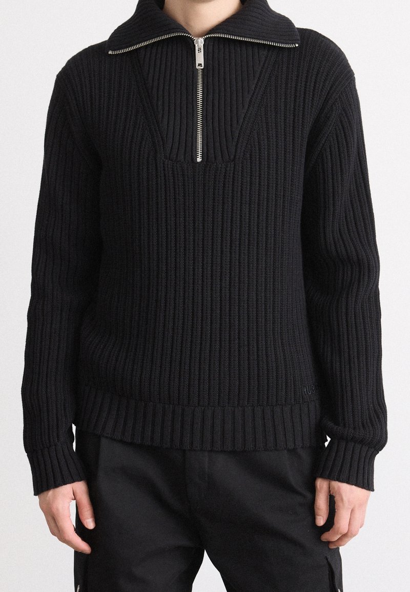 Personne portant un pull noir en maille côtelée avec col zippé mi-haut et manches longues, associé à un pantalon noir.