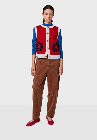 Crvena pletena prsluk s crnim cvjetnim uzorcima, bež rubom i zlatnim gumbe, nosi se preko plave majice i uparuje s smešnim čeurkanim hlačama i crvenim balerinkama.