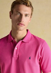 Roosa polo särk tekstuuriga viimistlusega, kolme nupuga kaelus ja must logo tikand vasakul rinnal. Lihtne krae ja lühikesed varrukad.