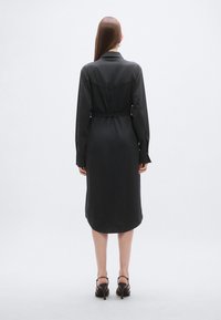 Μαύρο σατέν φόρεμα με γιακά, μακριά μανίκια και δεμένη μέση. Το μήκος είναι μέτριο, με ομαλή υφή και ελαφρώς καμπυλωτή τελείωμα.