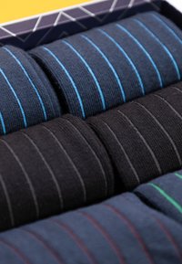 Vijf paar sokken in een doos, met donkere kleuren en verschillende dunne strepen in blauw, groen en rood. Gebreide textuur en opgerolde vorm.