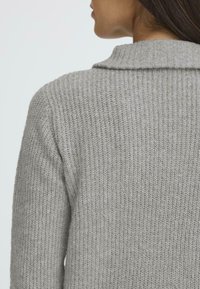 Pilkas megztinis su raštuota tekstūra, su sulankstyta apykakle ir laisvu kirpimu, matomas iš nugaros.