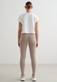 Femme avec un chignon tressé portant un crop top blanc, des leggings beige et des baskets blanches, debout face à un mur blanc uni.