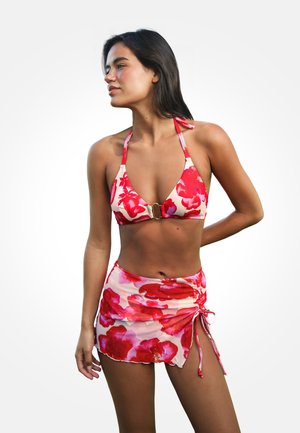 Woman wearing a red and pink floral bikini top with a matching sarong skirt, standing against a plain white background.