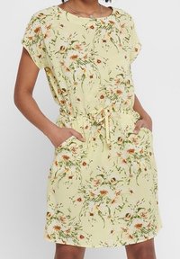 Robe fleurie en tissu jaune clair avec une texture douce. Comprend des manches courtes, une taille à cordon et des poches latérales. Motifs floraux verts et orange.