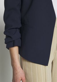 Navy blue blazer with rolled sleeves and a side pocket, paired with beige striped trousers, showcasing a textured fabric.