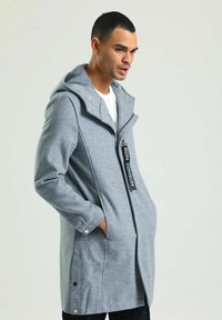 Grau Fleece-Mantel mit Kapuze, Frontreißverschluss, Seitentaschen und Logopatch. Besitzt einen leicht oversized Schnitt und Druckknöpfe an den Ärmelabschlüssen.