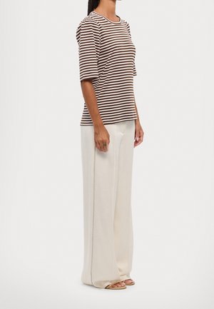 Kobieta ubrana w brązowo-biały pasiasty top z krótkim rękawem, szerokie białe spodnie i płaskie beżowe sandały, stojąca na jednolitym tle.