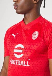 Maglia da calcio rossa con scollatura a V, caratterizzata da un motivo triangolare testurizzato, logo Puma bianco e stampa grafica che include la scritta "FOOTBALL".
