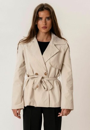 Hosszú barna hajú nő bézs, öves ballonkabátot visel fekete felső és fekete nadrág fölött, egyszínű világos háttér előtt.