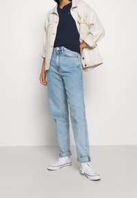 Světlé džíny z denimu ve volném střihu, doplněné tmavě modrým tričkem a krémovo-bílou denimovou bundou. Bílé sneakers dotvářejí vzhled.