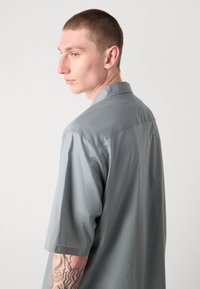 Camisa de manga corta con cuello en tela gris claro de textura suave. Presenta un corte relajado y un diseño con yugo trasero.