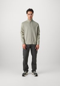 Világoszöld, zipzárral záródó pulóver, texturált mintával, sötétszürke cargo nadrággal és sötét sneaker cipővel, fehér talppal párosítva.