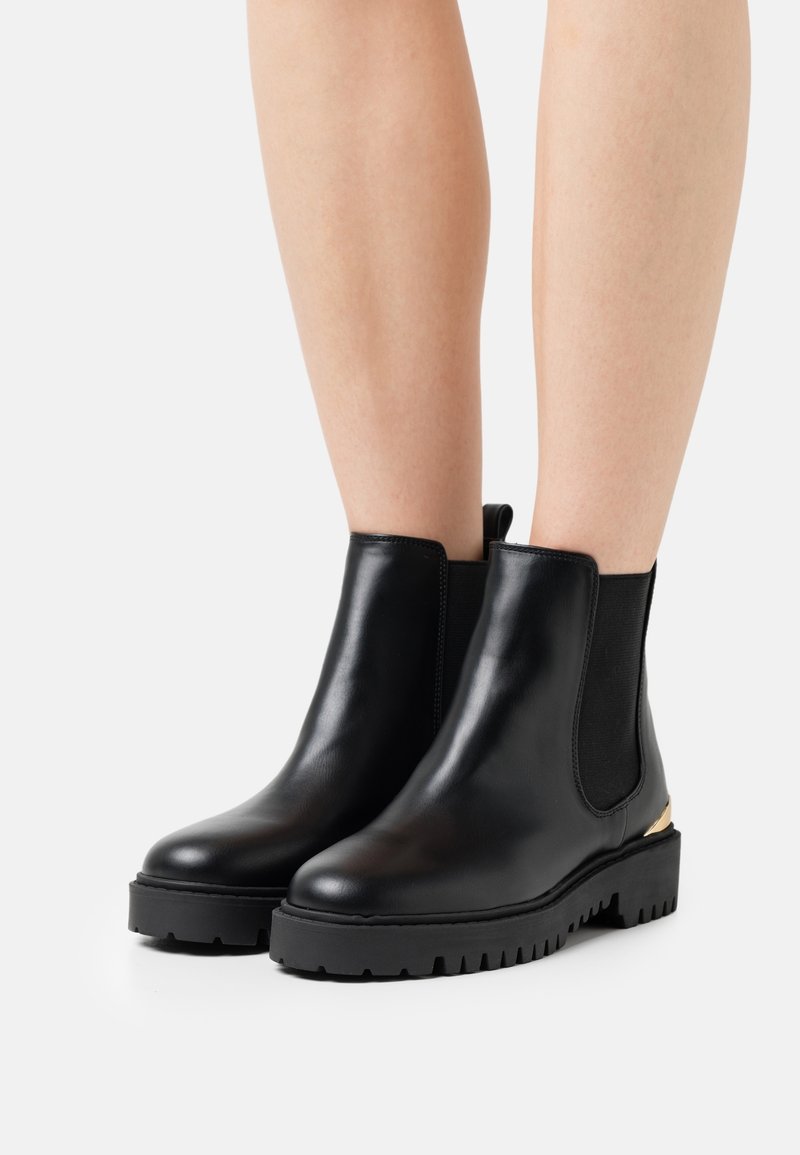 Bottines en cuir noir avec semelles en caoutchouc épaisses et panneaux latéraux élastiques, portées sur des jambes nues contre un fond blanc.