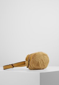 Kulatá hnědá kožichová ledvinka s koženým popruhem a stříbrným zipem umístěná na bílé podstavě.