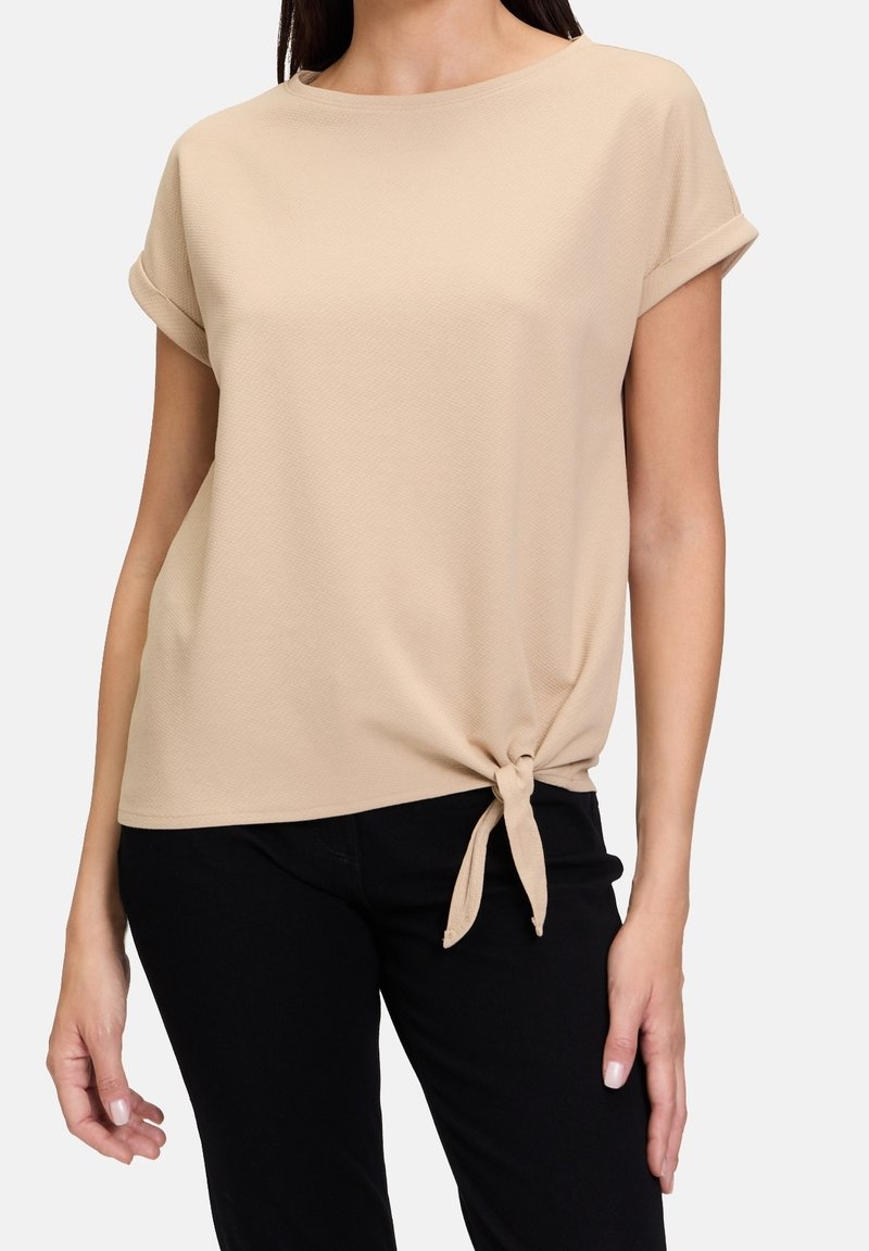 Beige top s krátkým rukávem, kulatým výstřihem a texturovaným materiálem. Uzlový detail v pase. V kombinaci s černými kalhotami.