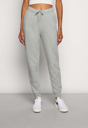 Vrouw draagt lichtgrijze Nike joggingbroek met tunnelkoord in de taille en witte Nike sneakers, tegen een effen achtergrond.