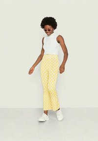 Top sem mangas branco com textura canalizada, combinado com calças largas amarelas com padrão de xadrez. Modelo usa óculos de sol redondos e tênis brancos.