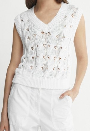 Donna che indossa un gilet bianco senza maniche lavorato a maglia con motivo a treccia a V con dettagli in mesh e pantaloni bianchi, mano in tasca.
