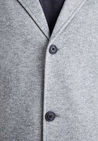 Primo piano di un cappotto di lana grigio con due bottoni neri allacciati sopra un indumento interno grigio scuro.