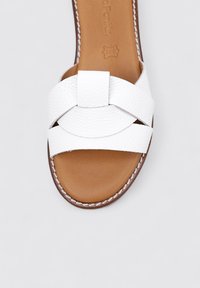 Sándala de piel blanca con una tira ancha de diseño texturizado, un acento circular y una plantilla color marrón con costuras visibles.