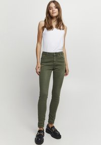 Jeans ajustados verde oliva con una textura suave, cierre de botón y bolsillos frontales, combinados con una blusa sin mangas blanca y zapatos de plataforma negros.