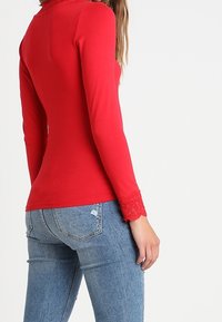 Maglietta rossa a maniche lunghe realizzata in tessuto elasticizzato, con dettagli in pizzo ai polsini e una silhouette aderente, abbinata a jeans in denim blu.