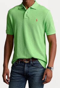 Grön polo skjorta i texturerad tyg, med en tre-knapps placket och en liten röd logotyp på bröstet, i kombination med mörka jeans.