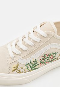 Baskets en toile beige avec lacets blancs. Présentent une broderie florale en vert, rouge et jaune sur les côtés. Semelle en caoutchouc.