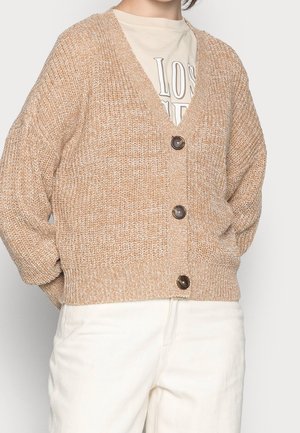 Persoon die een beige grofgebreid vest met drie knopen draagt over een crèmekleurig t-shirt met print en een witte broek, met de handen in de zakken.