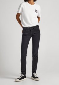 Weißes T-Shirt mit schwarzem "Pepe Jeans" Logo, kombiniert mit eng anliegenden schwarzen Jeans und schwarzen High-Top-Sneakern mit weißen Sohlen.