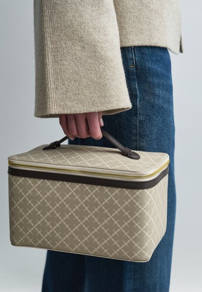 Μπεζ υφασμάτινη τσάντα με γεωμετρικό σχέδιο, καφέ λεπτομέρειες, χειρολαβή στην κορυφή και κίτρινο φερμουάρ, που κρατιέται σε χέρι κοντά σε μπλε τζιν.