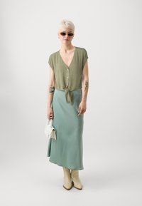 Blusa corta a maniche corte color verde oliva con nodo frontale, abbinata a una gonna fluida color verde menta. Borsa bianca e stivali da cowboy color crema.