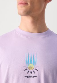 Svetlofialové bavlnené tričko s grafickým dizajnom, na ktorom sú modré hroty a žlté slnko pod textom "SCOTCH & SODA Amsterdam."