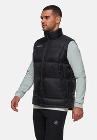 Schwarze gepolsterte Weste mit hohem Kragen und Frontreißverschluss, mit horizontalen Steppabschnitten und einer Seitentasche. Getragen über einem hellblauen Langarmshirt.