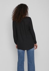 Maglietta nera a maniche lunghe con vestibilità ampia, orlo arrotondato e dettagli a pieghe sulla schiena, abbinata a jeans svasati blu chiaro.